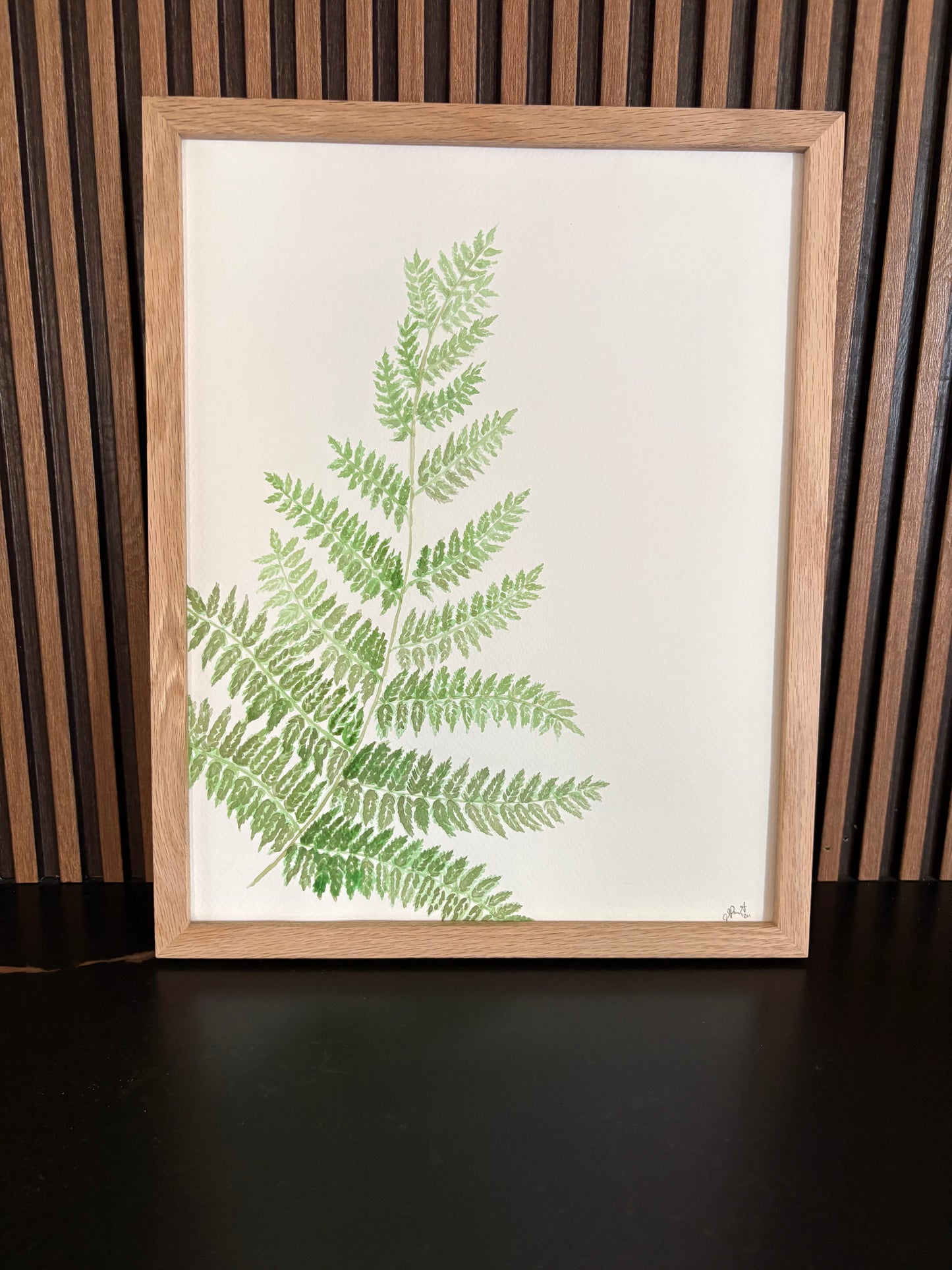 Framed artwork print of green fern leaves on a wooden slat wall on a black surface
