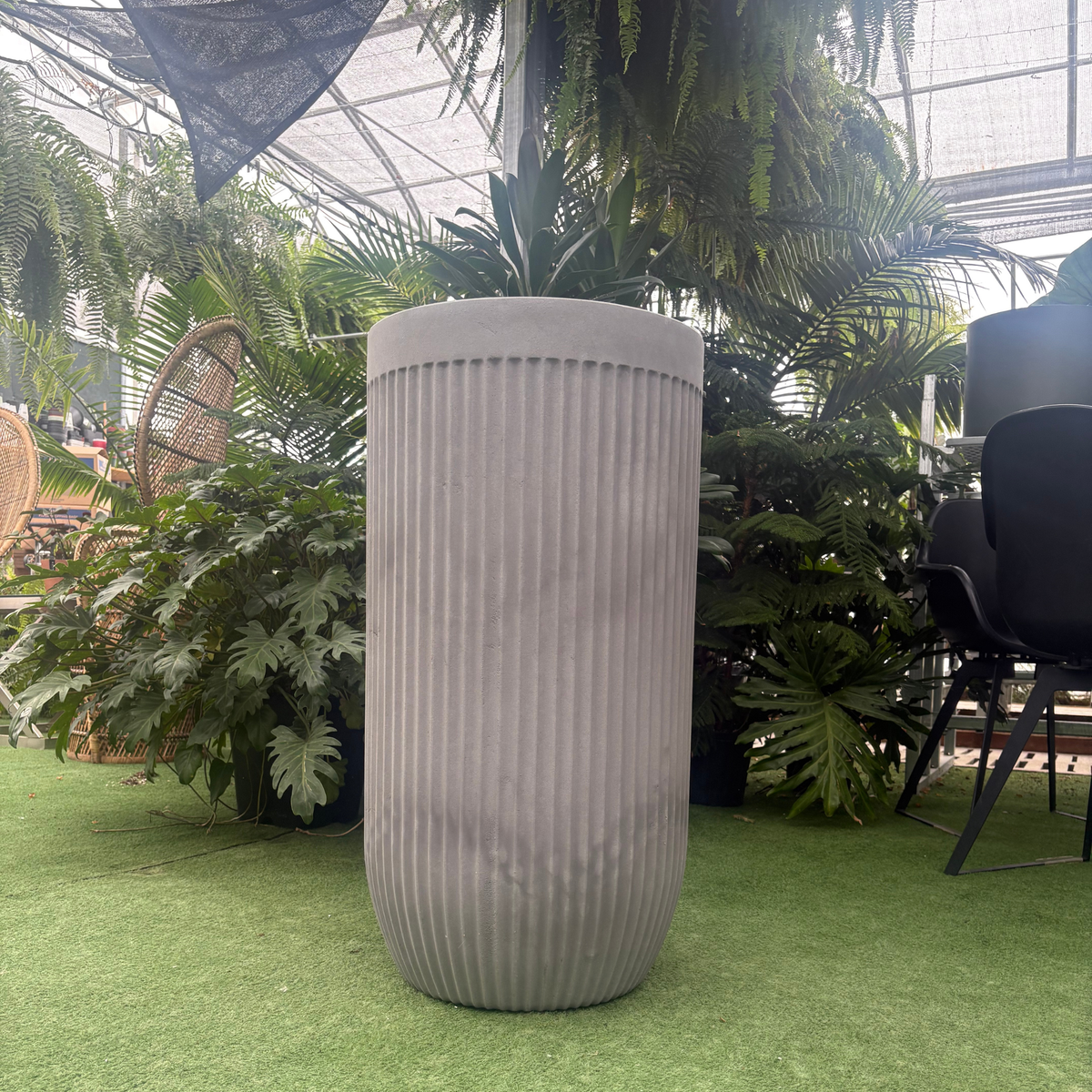 a burnt cement coloured flute round planter resting on a grass like setting in a greenhouse setting