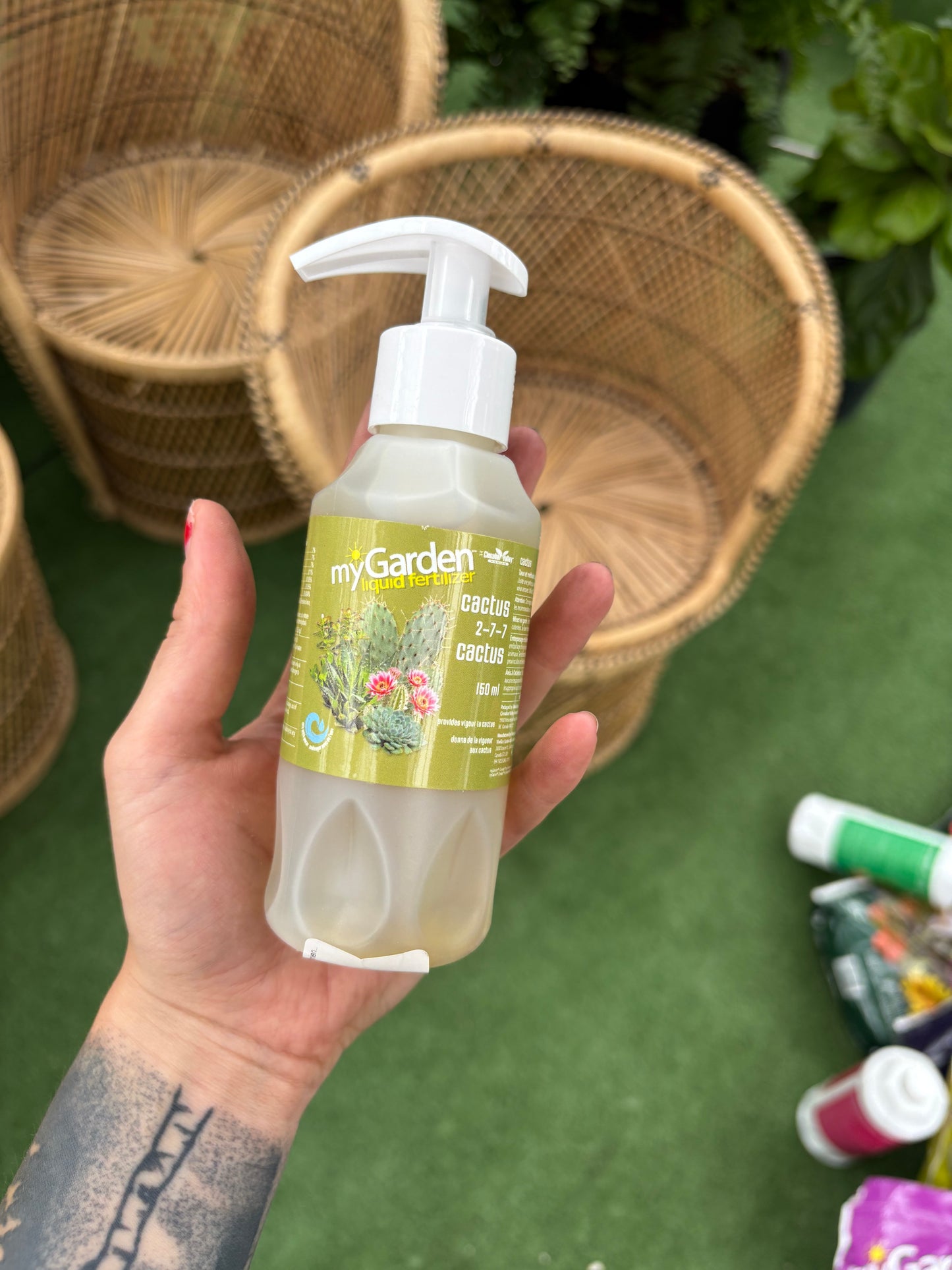 Hand holding a bottle of 'my garden' plant food with a wicker chair and green grass in the background.