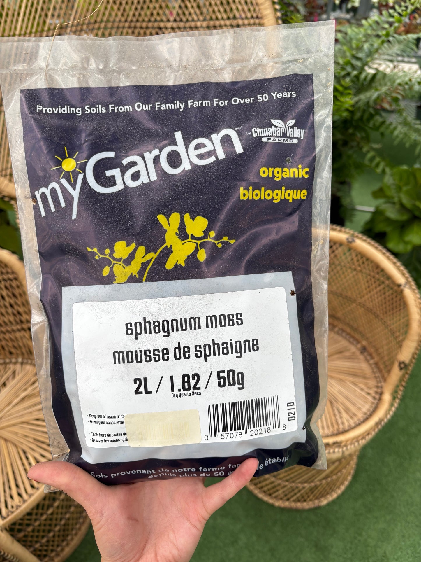 Hand holding a package of sphagnum moss labeled 'myGarden' with a wicker chair in the background.