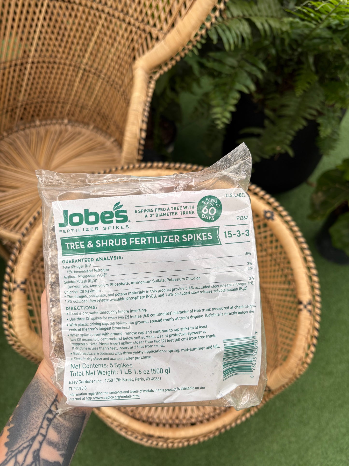 hand holding package of Tree & Shrub Fertilizer Spikes in front of wicker chairs and plants