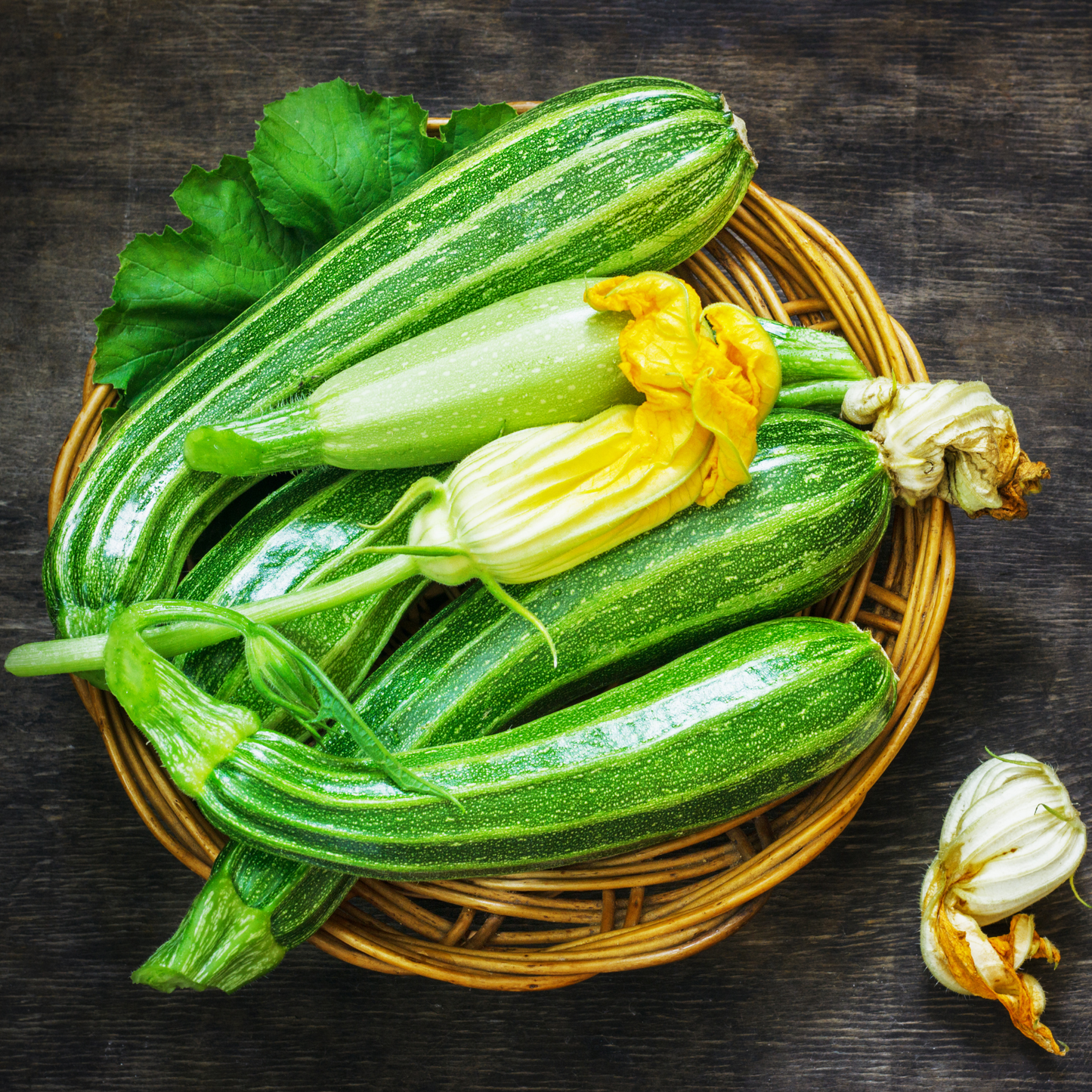 a wicker basket full of striped bright green flowering zucchini with yellow flowers against a wooden surface