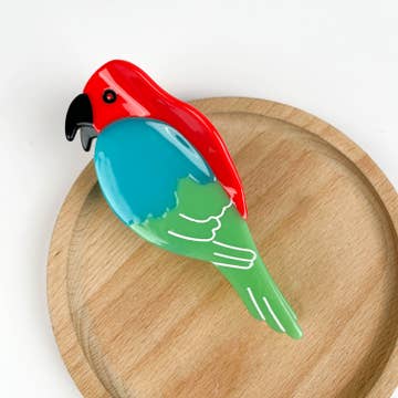 red, blue and green classic parrot hair clip resting on wooden disk with white background