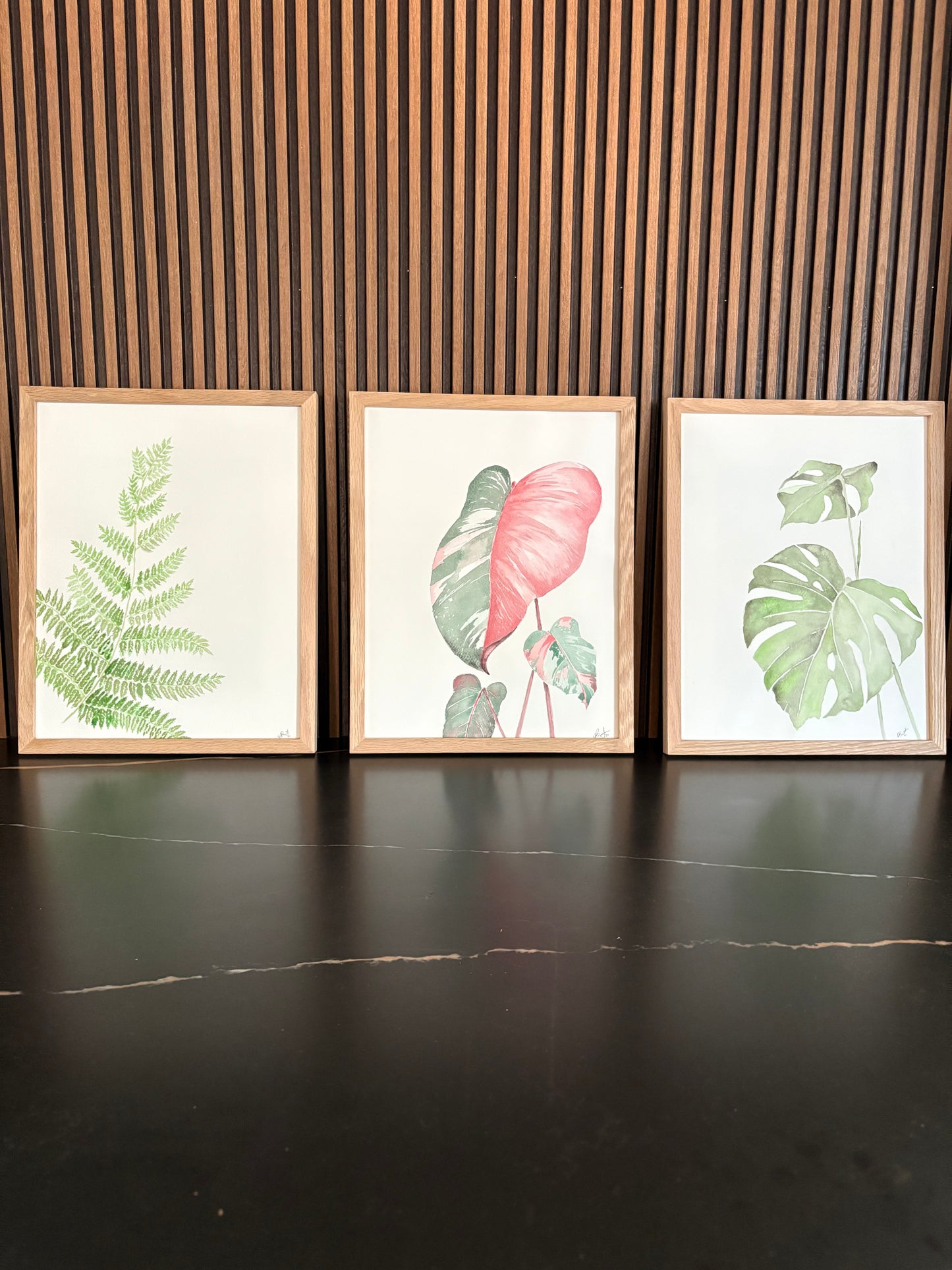 Three framed botanical prints on a wall with wooden horizontal slats on black surface