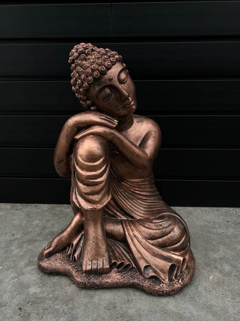 Bronze Buddha statue with arms and head resting upon one knee, the statue is sitting on concrete floor with black slate background