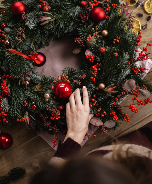 Decorative Christmas wreath with red ornaments and a hand reaching towards it on a wooden surface.