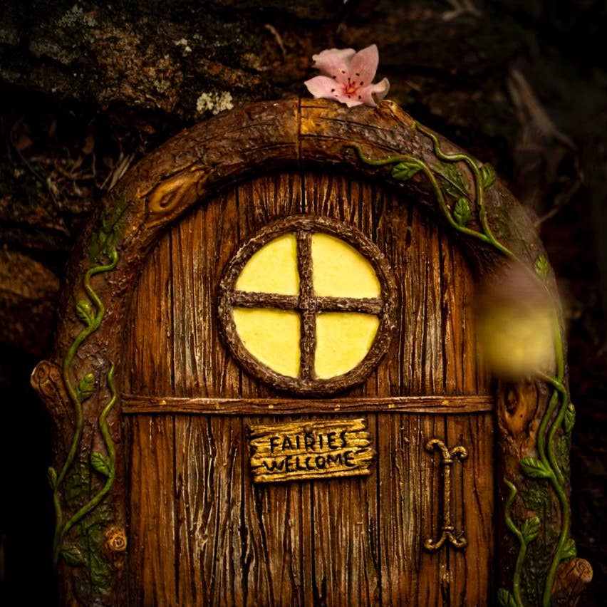 A whimsical wooden fairy door with a round window and a sign that reads 'Fairies Welcome'. It is adorned with a green vines around the door frame and a pink flower is visible on the left side.
