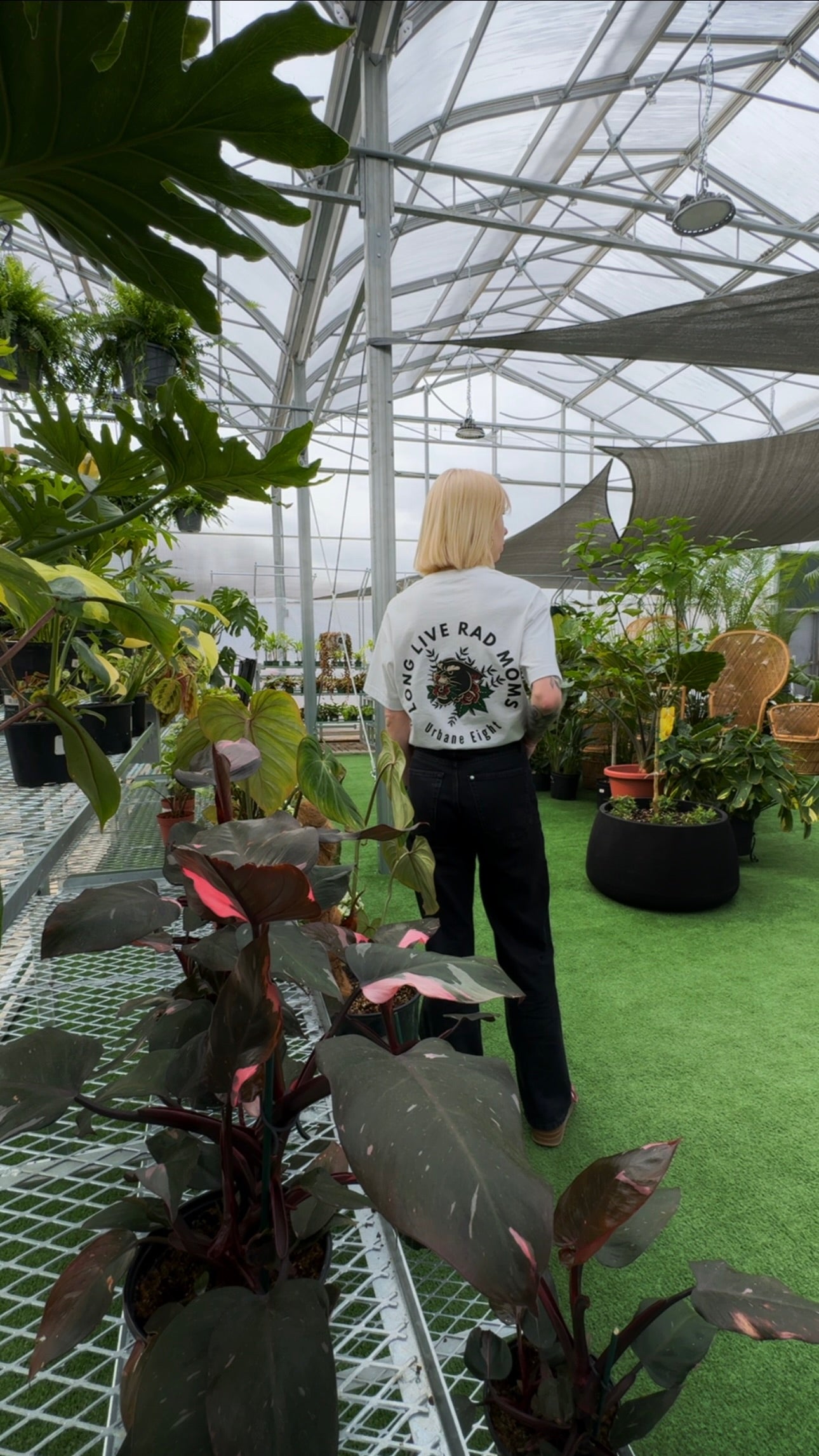 a woman standing in a greenhouse setting waring a white t-shirt that says long live rad moms