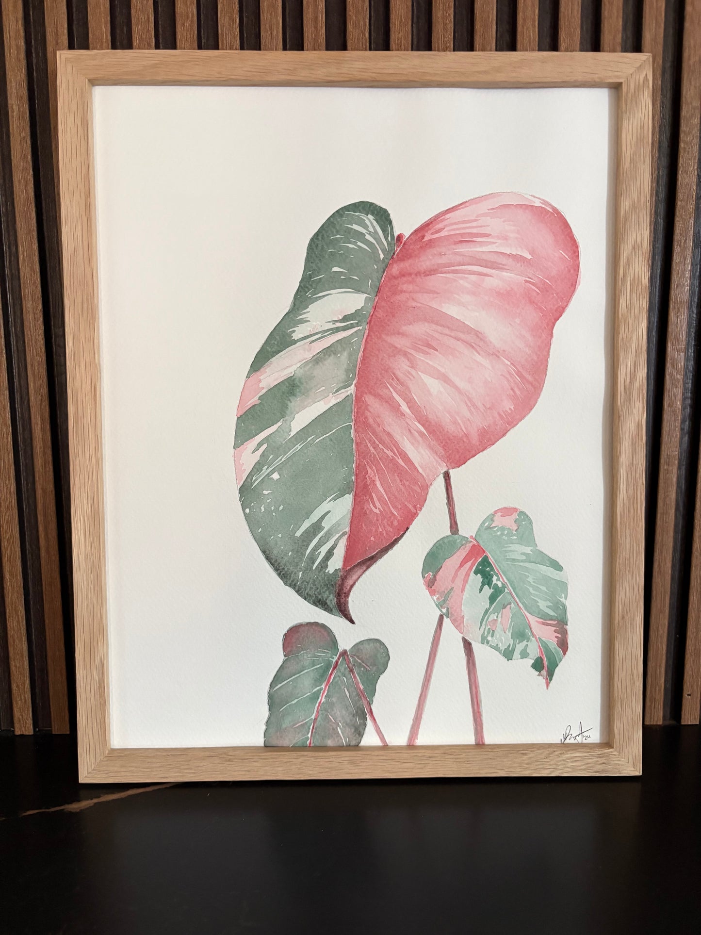 Framed artwork of pink and green philodendron leaves on a wooden slat wall with a black surface
