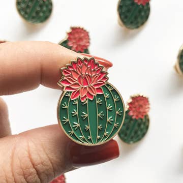 a collection of cactus shaped pins with green plant and pink flowers held by a hand against a white background