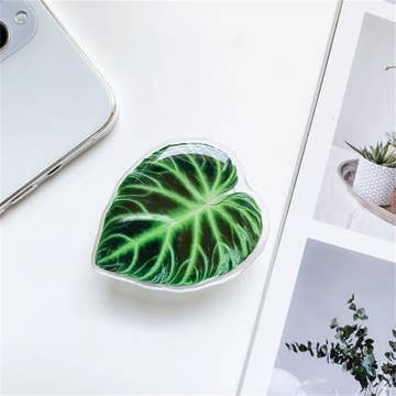 A heart-shaped, green plant-themed phone holder placed on a surface next to a mobile phone and a plant pot.