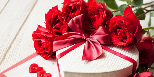 a heart shaped white gift box wrapped with a red bow surrounded by red roses