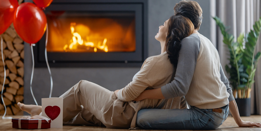 a couple cuddling by the fire with a heart shaped card and red balloons beside them with plants in the background