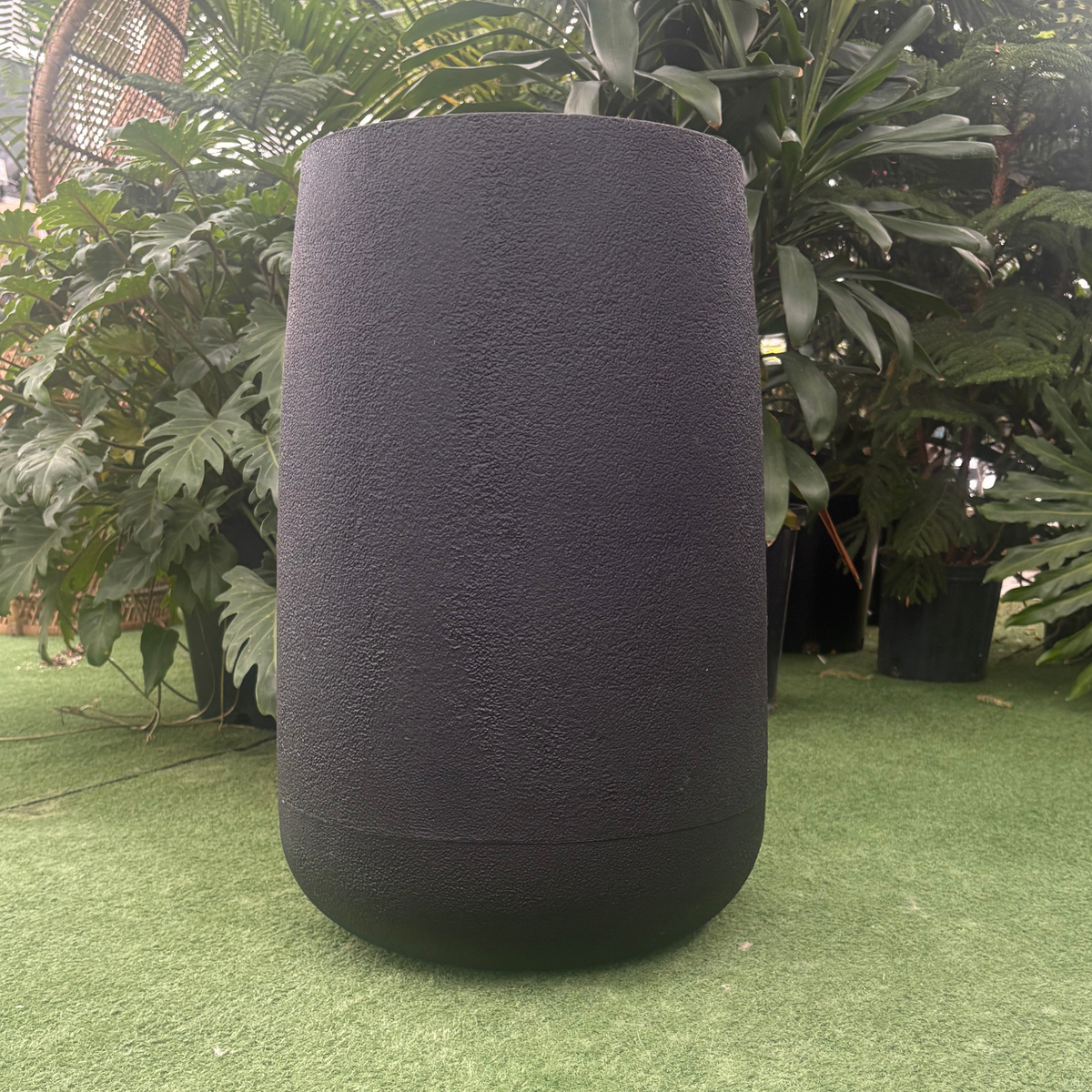 a close up of a round edged matte black textured bios round planter resting on a grass like surface in a greenhouse setting