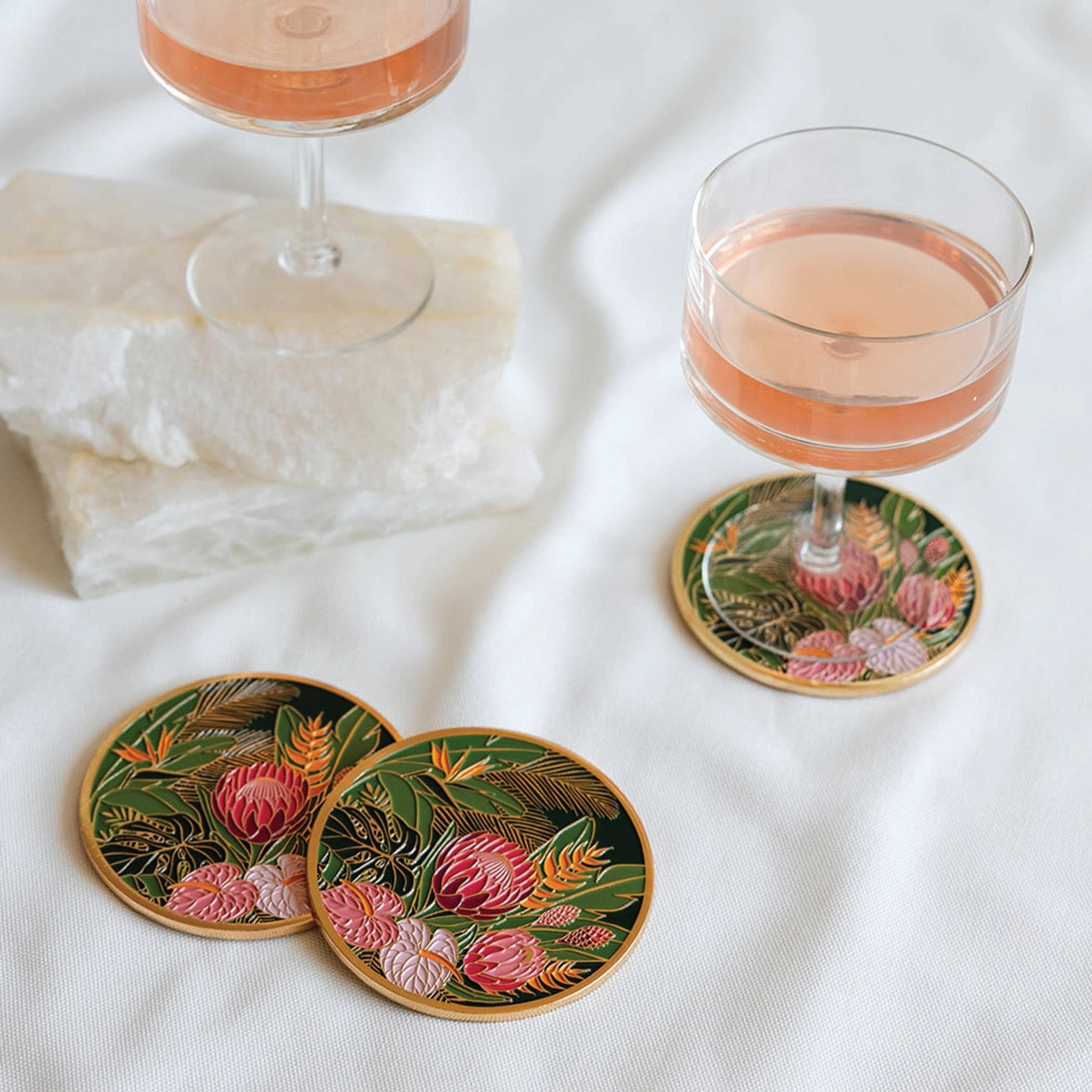 Two glasses of pink cocktail on floral coasters with a white napkin on a white surface