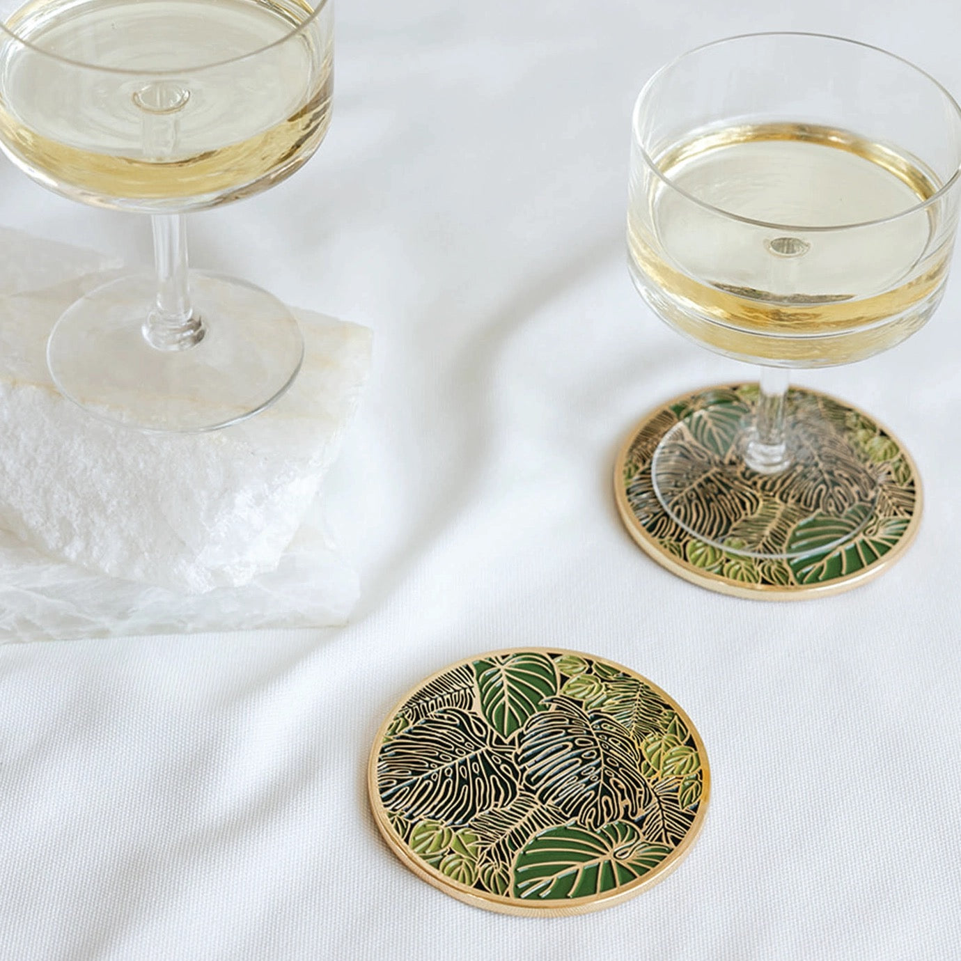 Two glasses of white wine on decorative coasters with leaf patterns on a white surface.