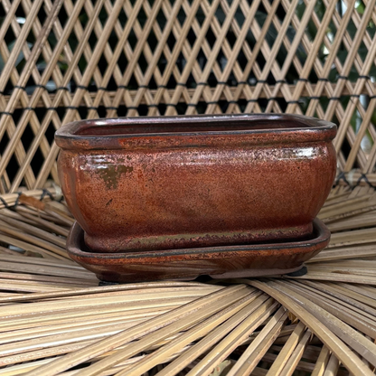 a copper toned rectangle shaped Chinese glazed iron ceramic bonsai pot with a drip tray resting on a wicker chair
