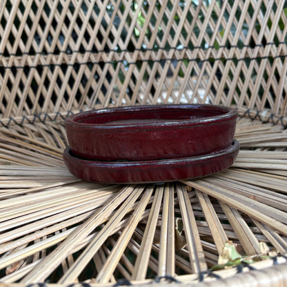 a small oval shaped dark red Chinese glazed oxblood ceramic bonsai pot with a drip tray resting on a whicker chair