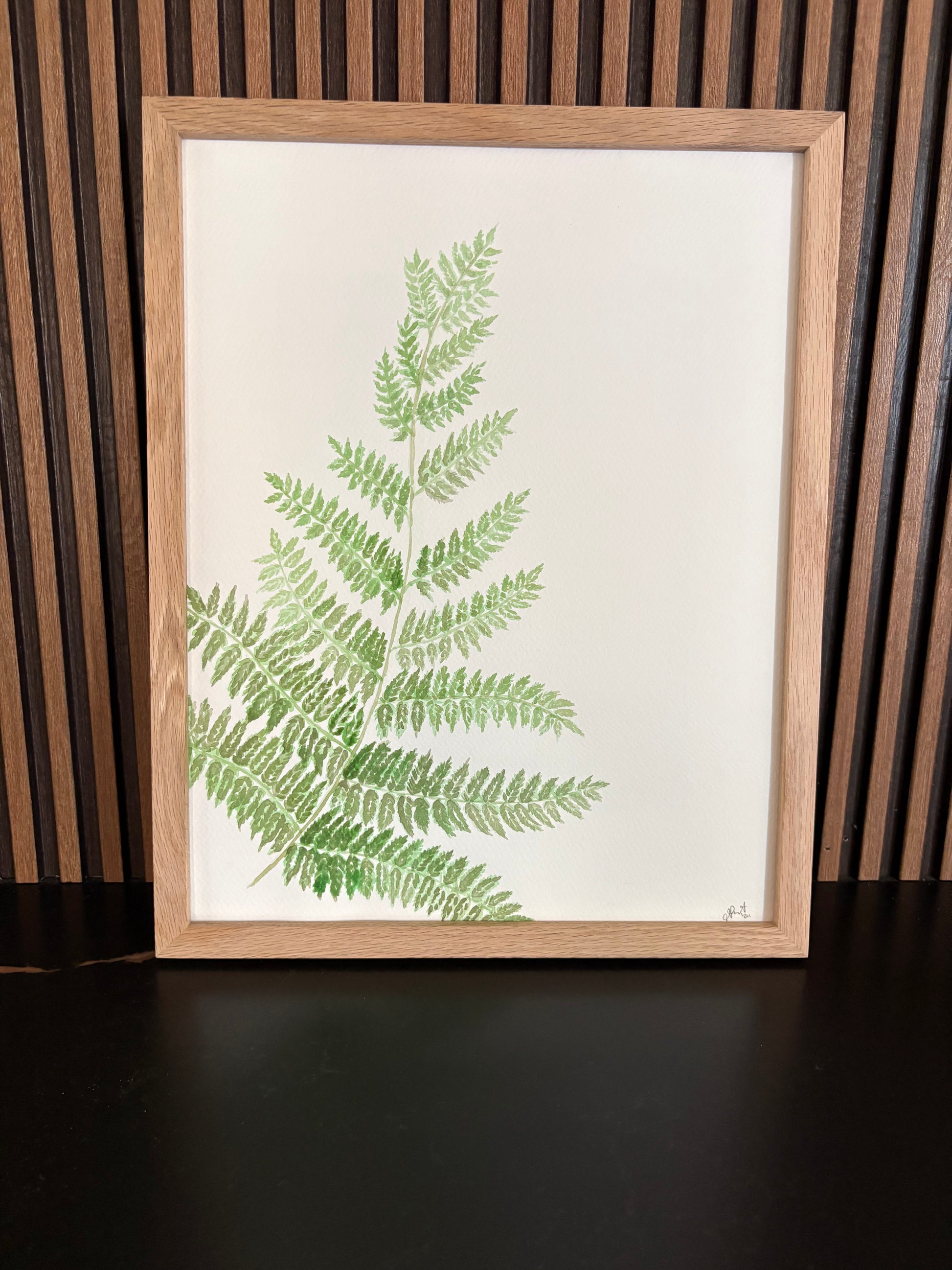 Framed artwork print of green fern leaves on a wooden slat wall on a black surface