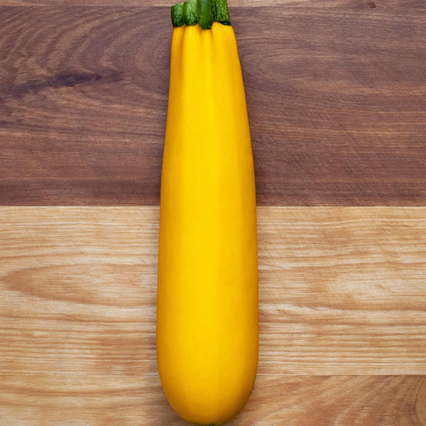 a bright yellow golden zebra zucchini with a green top resting on a wooden surface