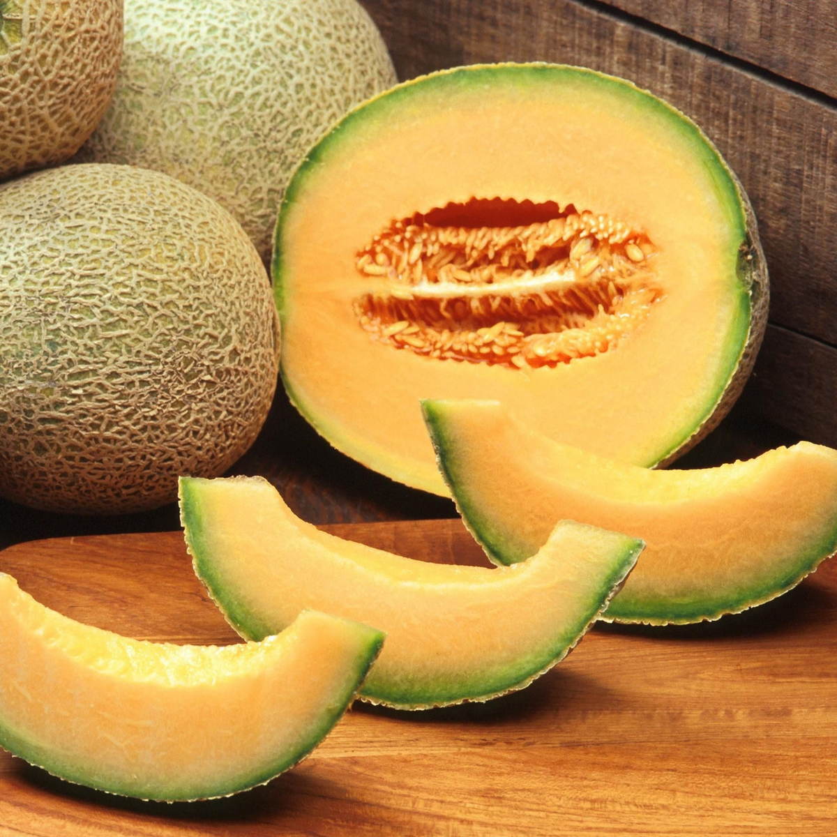 Fresh jumbo cantaloupe melon slices and a cut melon half displayed on a wooden surface, with whole melons in the background.