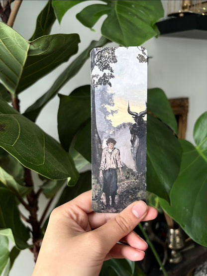 Hand holding a decorative bookmark with a Laura Alexander fantasy painting printed on it in front of green leaves.