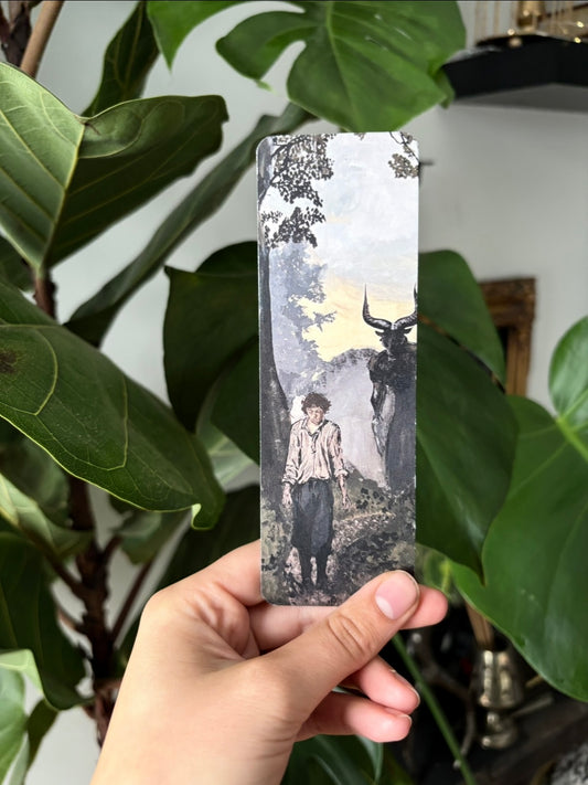 Hand holding a decorative bookmark with a Laura Alexander fantasy painting printed on it in front of green leaves.