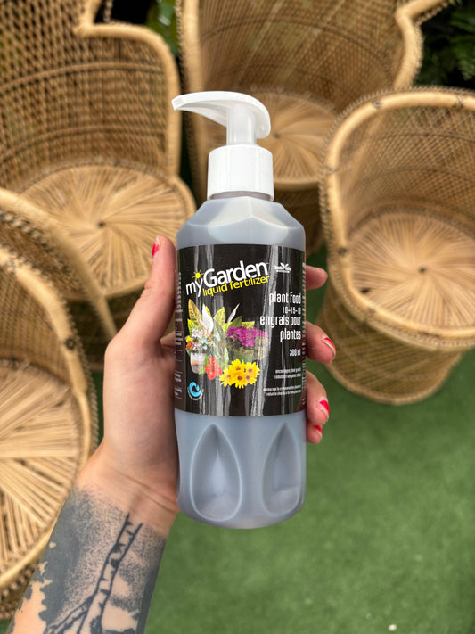 Hand holding a bottle of 'myGarden' plant food with wicker chairs in the background