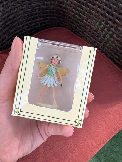 Hand holding a small Mini Flower Fairies figurine in a clear plastic case on a textured surface.