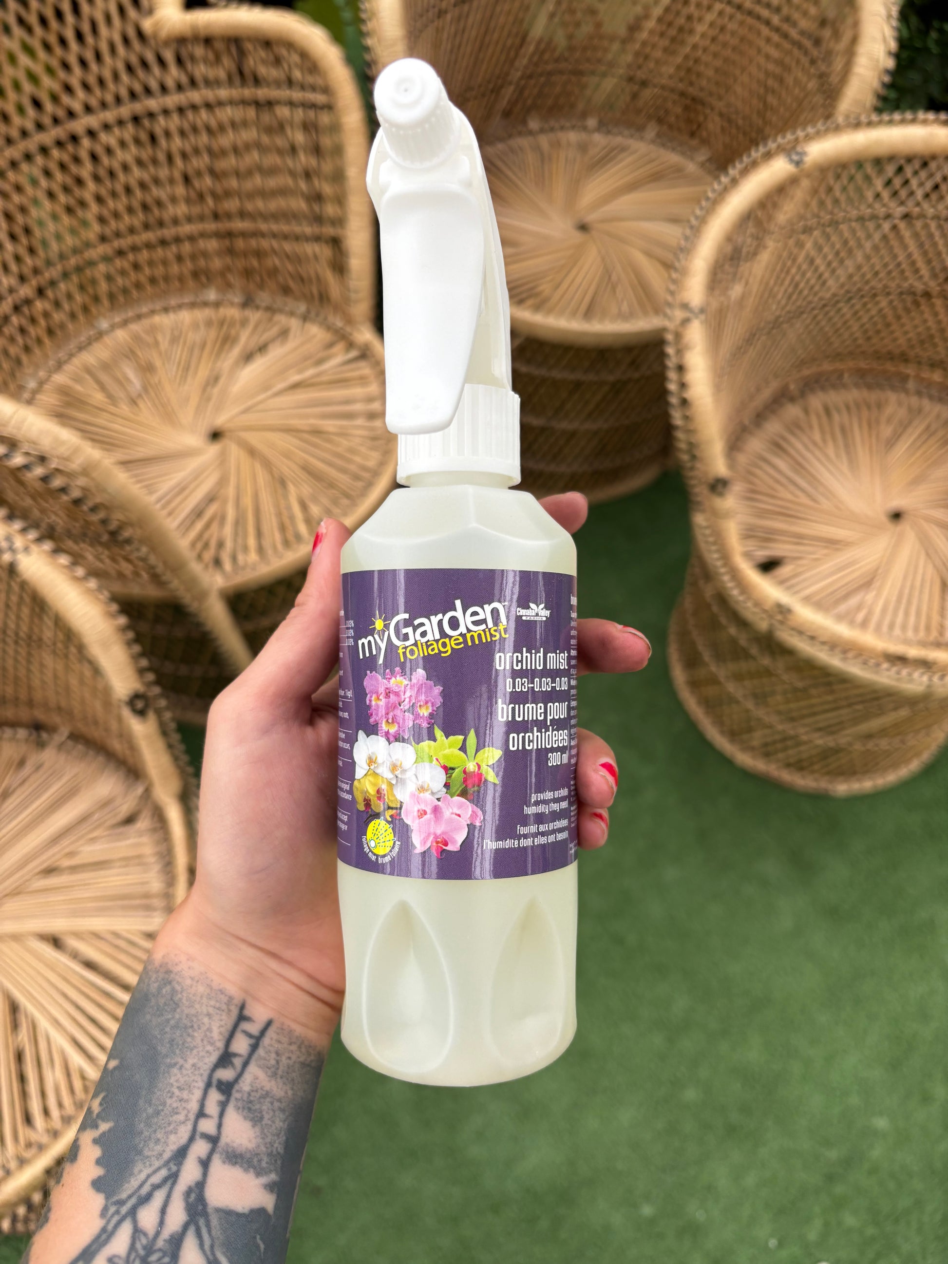 Hand holding a bottle of 'My Garden' plant food with wicker chairs in the background.