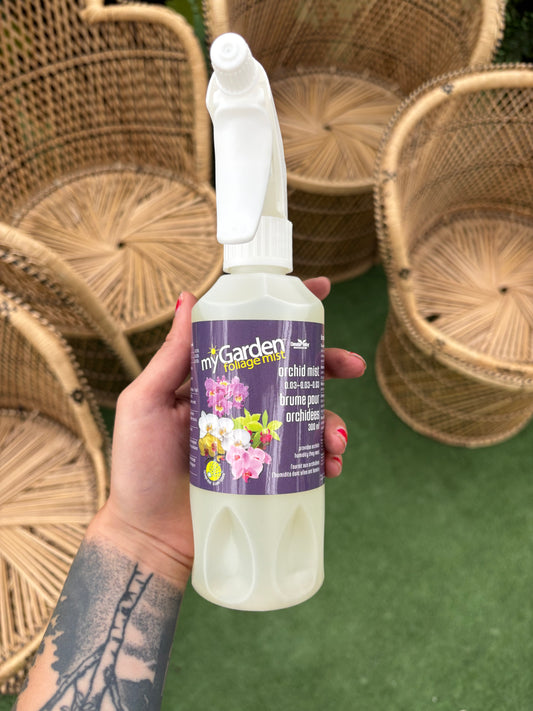 Hand holding a bottle of 'My Garden' plant food with wicker chairs in the background.