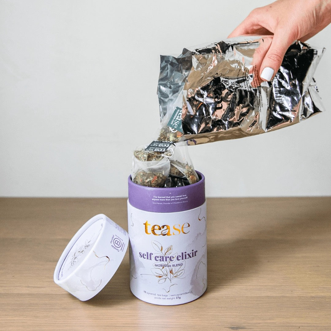 Hand pouring a package of 'tease' self-care elixir refill into the tea container on a wooden surface.