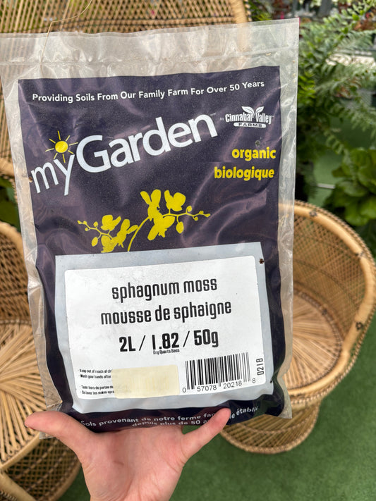 Hand holding a package of sphagnum moss labeled 'myGarden' with a wicker chair in the background.