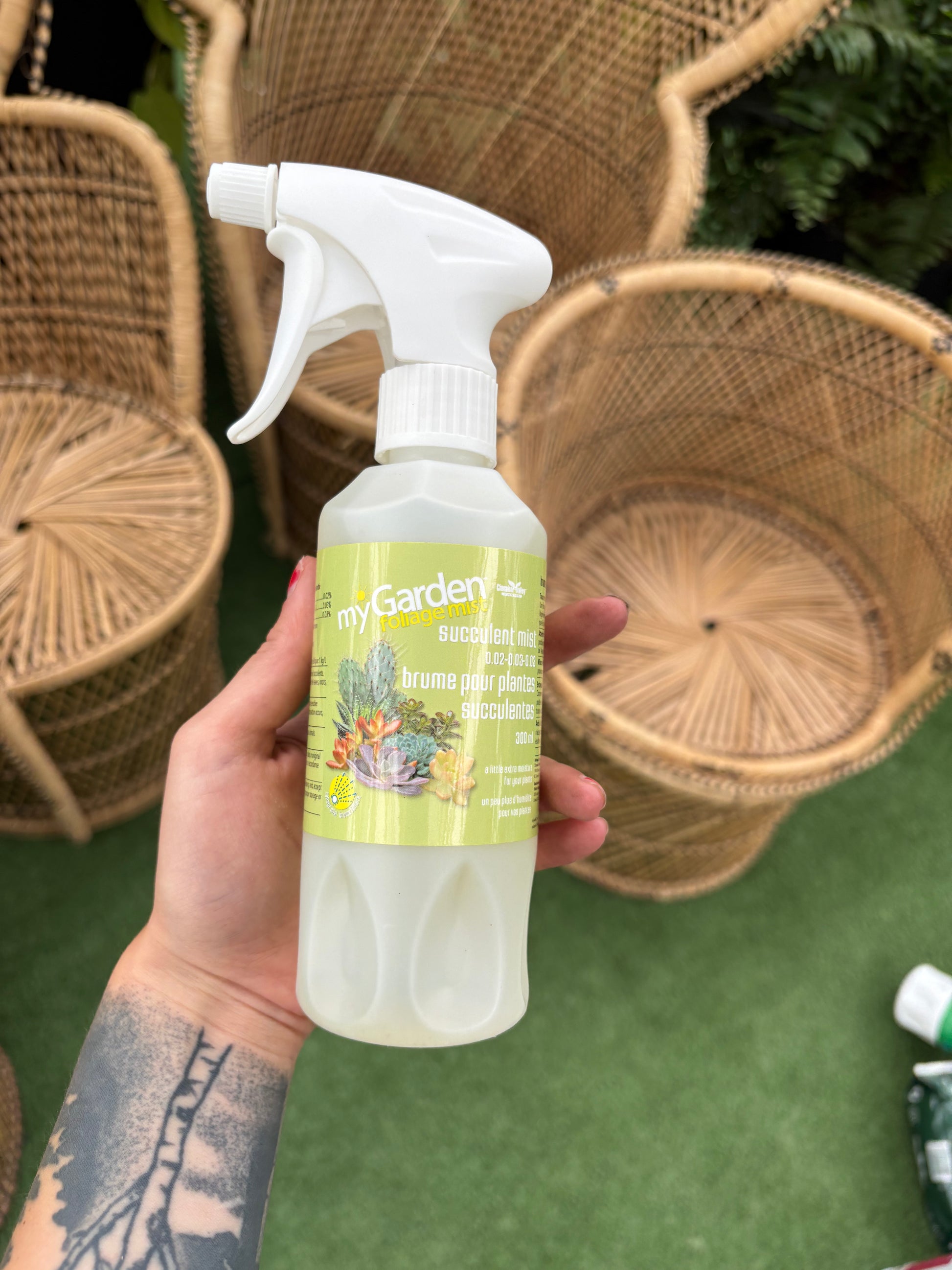 Hand holding a bottle of 'My Garden' plant food spray with wicker chairs in the background.