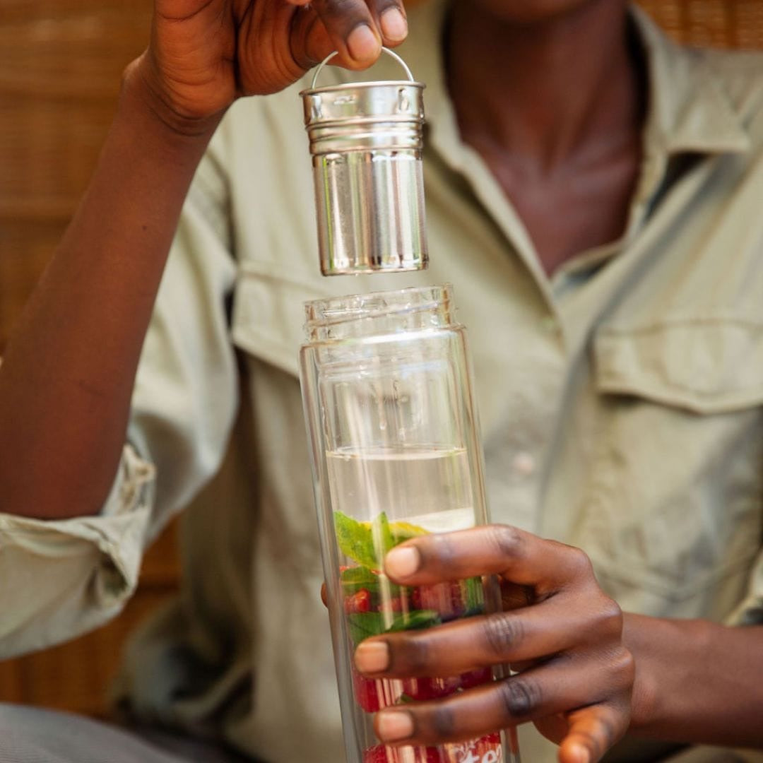 Person holding a clear bottle with a silver tea strainer lid, containing a red liquid and green leaves.