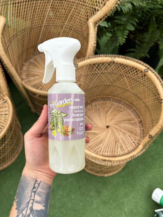 Hand holding a bottle of 'My Garden' plant food with wicker chairs in the background.