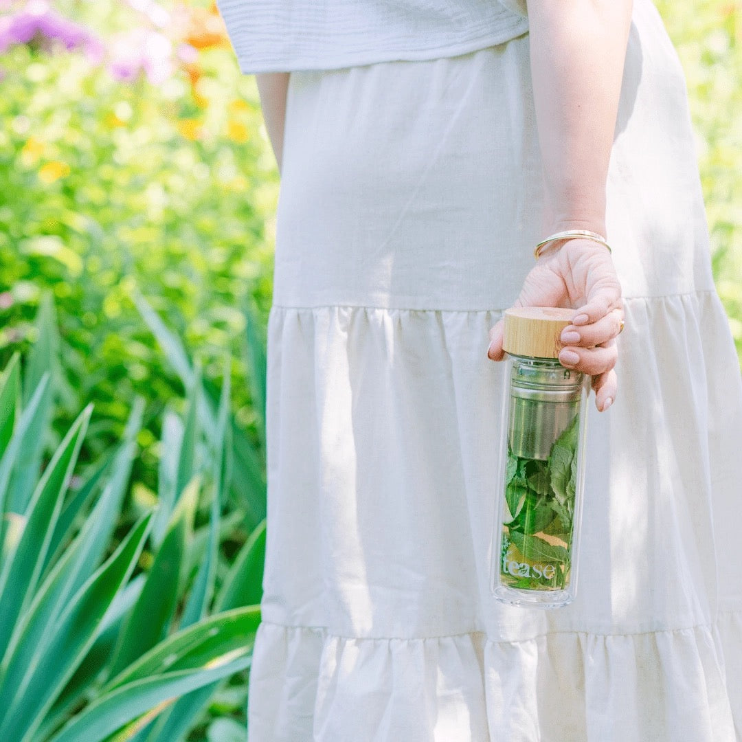Person holding a glass bottle with a wooden cap, filled with green leaves, against a blurred natural background.