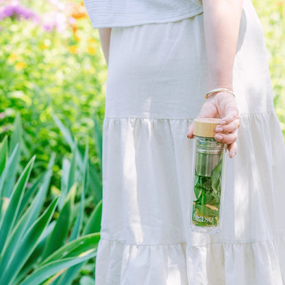 Person holding a glass bottle with a wooden cap, filled with green leaves, against a blurred natural background.