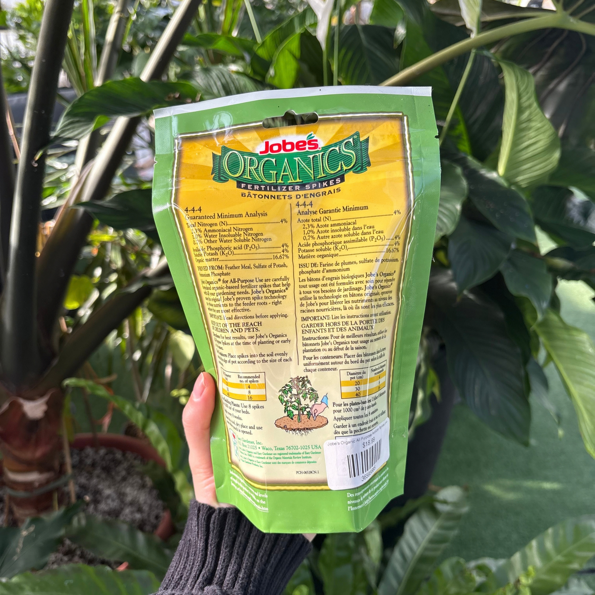 a person holding a package of jobs organics fertilizer spikes showing the back of the package that displays the ingredients and use instructions with many tropical plants in the background