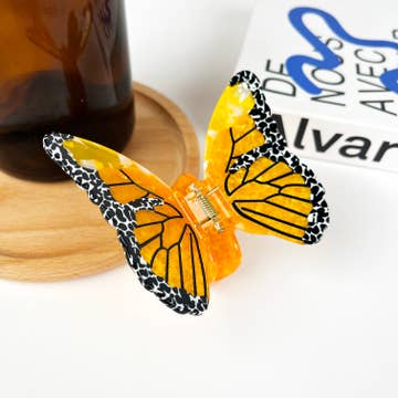 A butterfly-shaped hair clip with black and orange wings, resting on a wooden surface next to a glass and a book.