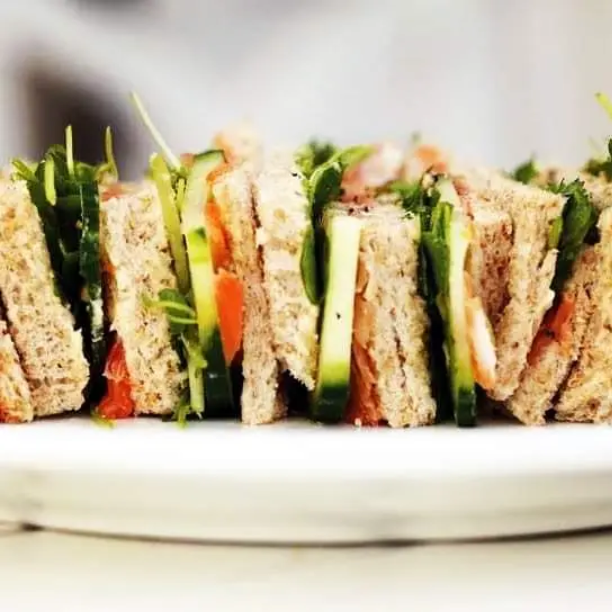 A close-up of a sandwich spread filled with vegetables and basil micro greens on a white plate, featuring whole wheat bread, cucumber, tomato, and greens.