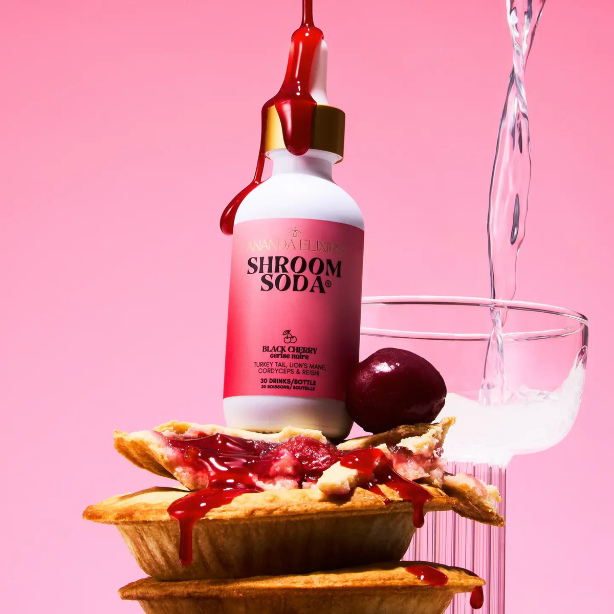 a white dropper bottle with a dark pink label that reads shroom soda black cherry with the bottle standing on multiple small cherry pies and a beverage being poured behind it with a pink background