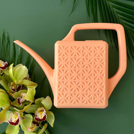 A matte peach tone watering can with breeze block-inspired geometric detailing on the side, displayed next to some green plants.