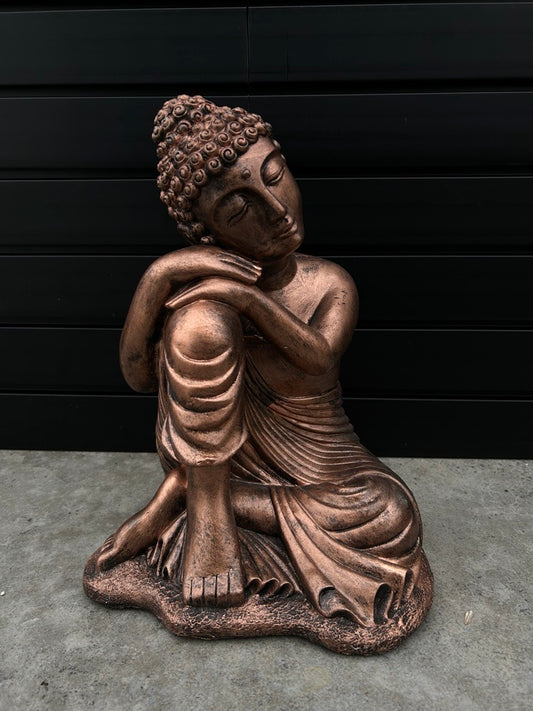 Bronze Buddha statue with arms and head resting upon one knee, the statue is sitting on concrete floor with black slate background