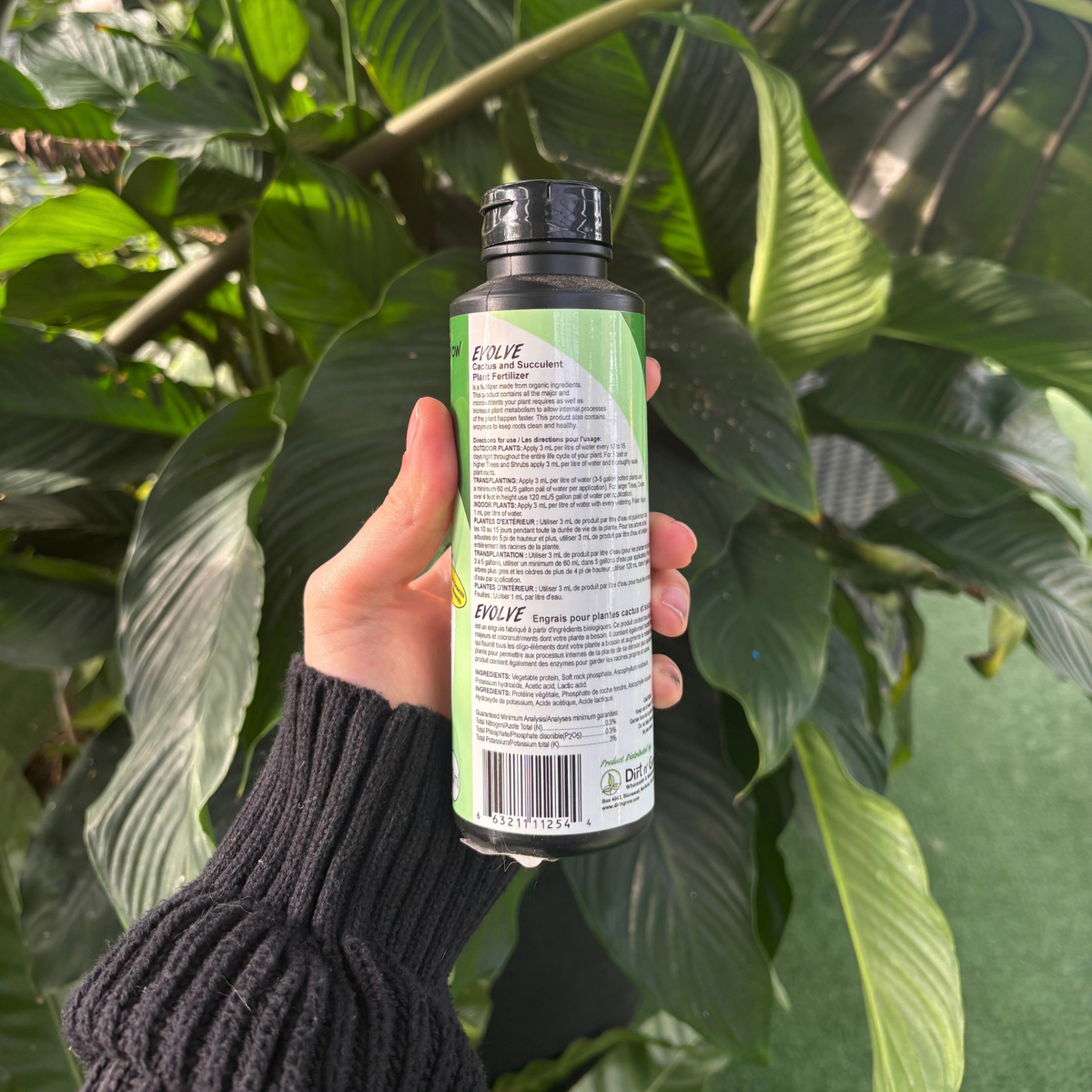a person holding a black bottle showing the back of the product with the label that indicates the ingredients and use instructions with many tropical plants in the background