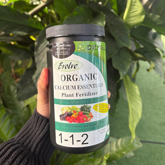 a person holding a black container with a label that says organic calcium essentials plant fertilizer with many tropical plants in the background
