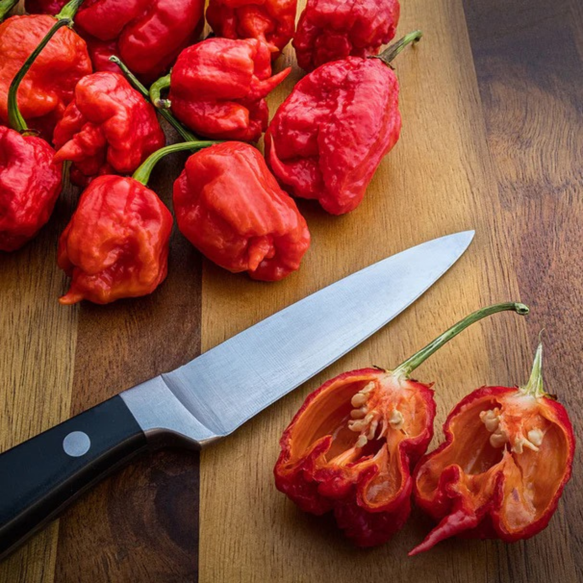 a pile of bright red Carolina reaper peppers next to a knife and one pepper cut open on a cutting board