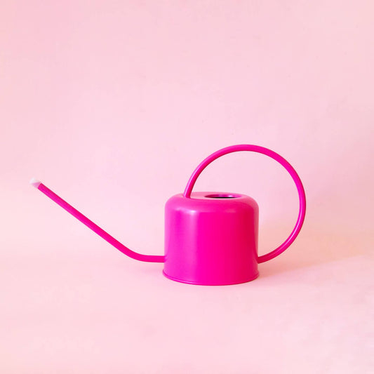 A vibrant hibiscus pink metal watering can with a long, slender spout, displayed against a pink background.
