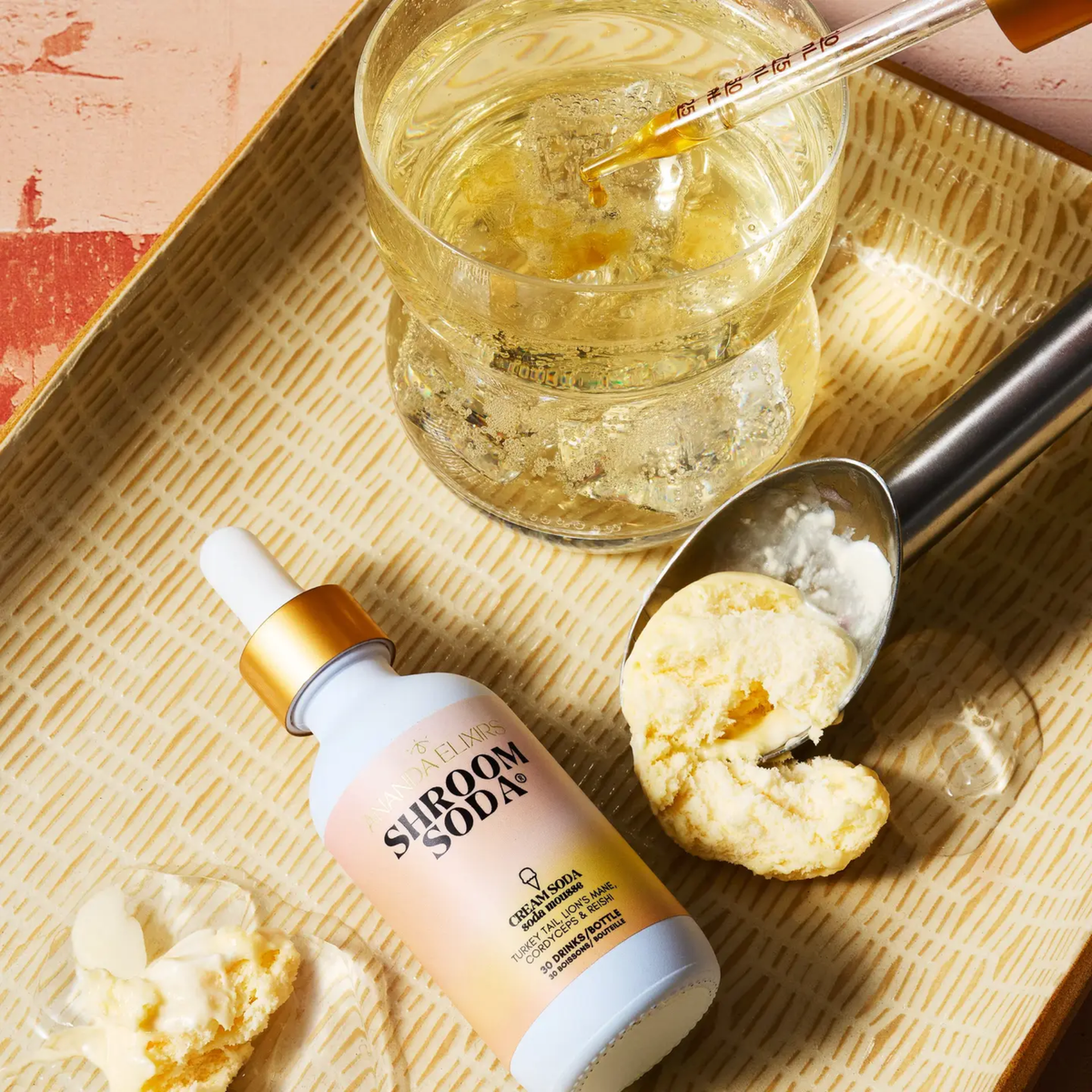 a white dropper bottle with a pink and yellow label that says shroom soda cream soda, resting in a wicker basket ext to a scoop of ice cream and a sparkling beverage on a table