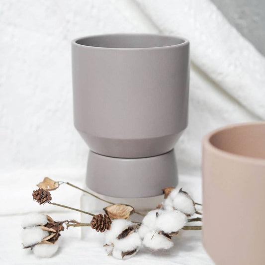 Gray ceramic cups with saucers, one larger than the other, displayed on a white textile surface. A few dried plant stalks in front of planter.